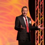 Shows a live event speaker on the stage speaking.